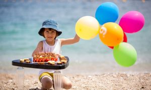 Young boy at the beach with balloons sitting with a birthday cake.