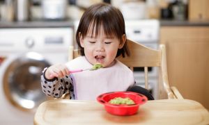 Baby in highchair eating avocado puree.