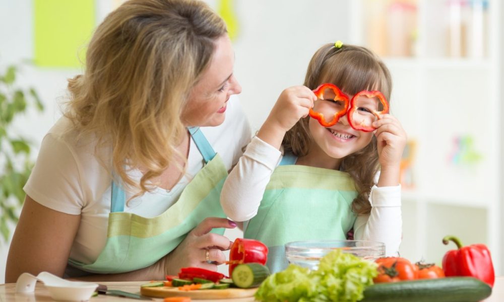 A mother and daughter cooking in the kitchen, with the child holding pepper slices up to her eyes.